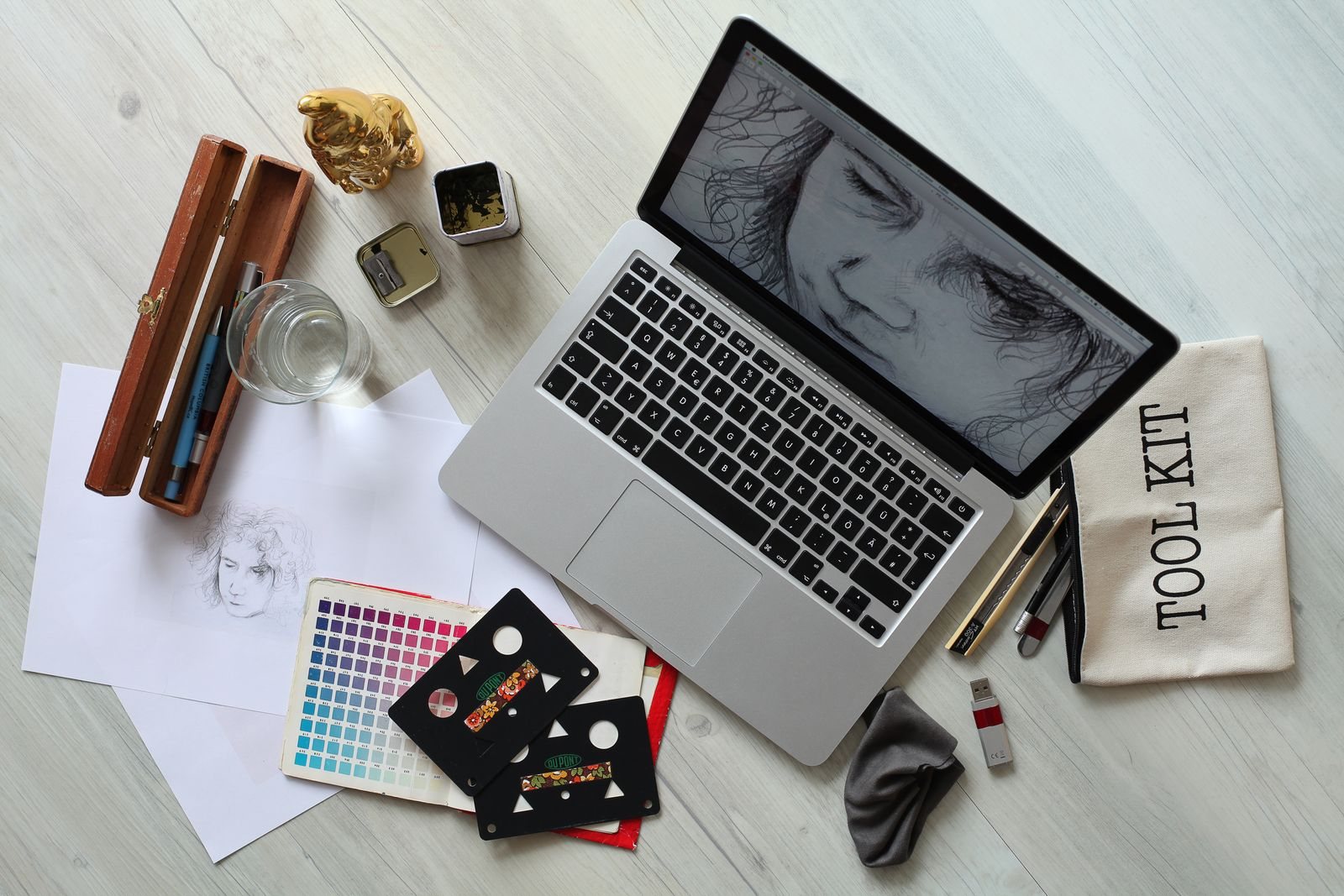 A laptop with a portrait sketch, colour swatches and pencils on a desk