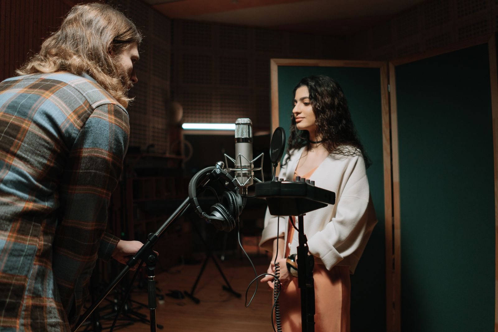 A singer at a microphone with a producer beside her in the vocal booth