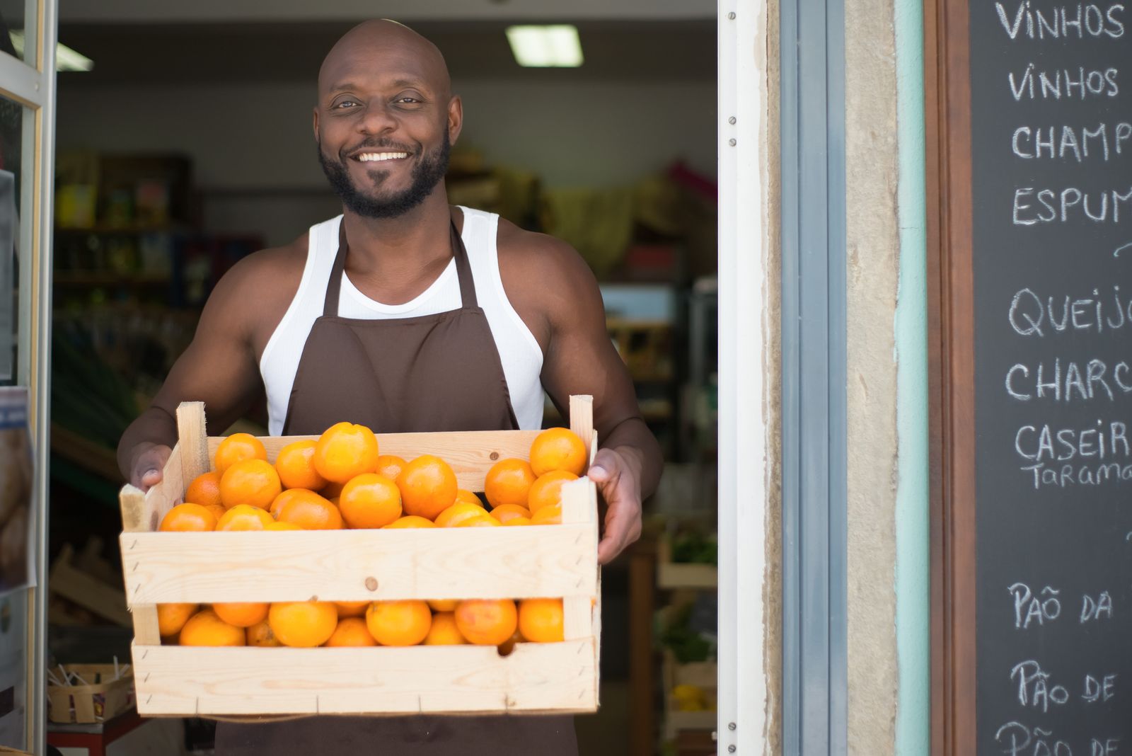 A grocer smiling in his shop doorway with a crate of oranges